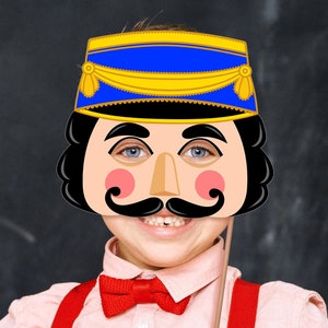 May include: A paper craft mask of a toy soldier with a blue hat, black hair, a pink face, and a brown moustache. The mask is on a stick and held by a child.