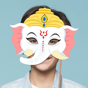 May include: A white paper mask with a pink elephant head and a yellow crown. The mask has red and black markings on the face and is held up to a person's face.
