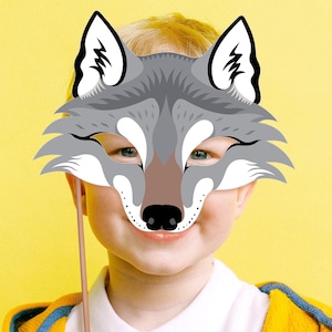 May include: A child holds a gray wolf mask in front of their face. The mask has a stick attached to it. The child is wearing a yellow shirt and is smiling.