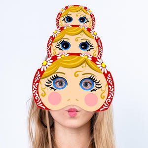Peut inclure: Trois masques de visage de poupées Matryoshka avec des cheveux blonds, des yeux bleus et des joues roses sont empilés. Les masques sont rouges avec des accents floraux blancs et sont tenus par un bâton en bois. L'adresse du site Web est visible en bas.