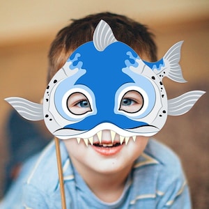 Könnte beinhalten: Ein Kind hält eine blaue und graue Fischmaske mit weißen Zähnen und Augen. Die Maske ist an einem Stock befestigt.