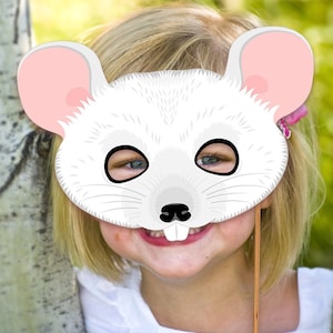 May include: A white mouse mask with pink ears and black eyes. The mask is held up to a child's face, who is smiling.
