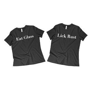 May include: Two black t-shirts with white text. The left shirt says "Eat Glass" and the right shirt says "Lick Rust".