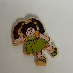 May include: A flat, die-cut plastic charm featuring a cartoon doll with dark brown hair in pigtails, wearing a green dress with yellow trim and matching green shoes. The doll has freckles, a round face, and is depicted in a dynamic pose.
