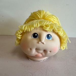 May include: A yellow yarn doll head with blue eyes and a pink mouth. The doll head is sitting on a white surface with a floral pattern.