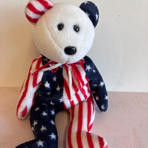 May include: A white plush bear with red and white stripes and blue stars on its body. The bear is wearing a white ribbon around its neck.