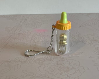 以下が含まれることがあります： 黄色い蓋と緑色の乳首が付いた、透明なプラスチック製の小さなベビーボトルキーホルダー。ボトルの中には、チェーンでつながれた金色のボールが入っています。