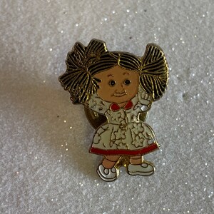 May include: A gold-colored pin featuring a cartoon girl with brown hair in pigtails, wearing a white dress with red trim and white shoes. The girl has a smiling face with brown eyes.