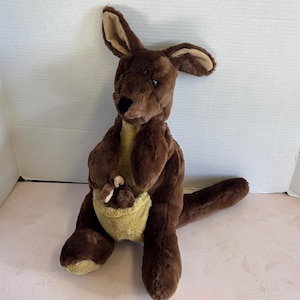May include: A plush kangaroo toy in shades of brown and tan. The kangaroo has large ears, a pouch, and a long tail. The toy is made of soft, fuzzy material and is designed to sit upright.
