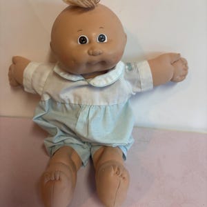 Peut inclure: Une poupée vintage pour bébé avec un teint hâlé, des yeux bruns et une touffe de cheveux blonds. La poupée porte une chemise à col blanc et un short à carreaux bleu clair. Les bras de la poupée sont tendus et elle a des pieds en tissu.
