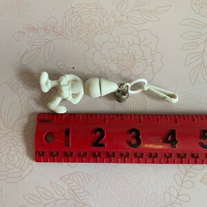 May include: White plastic keychain featuring a cartoon character with a large head and a small body. The character is holding a small object in its hand. The keychain has a metal ring and a plastic clip.