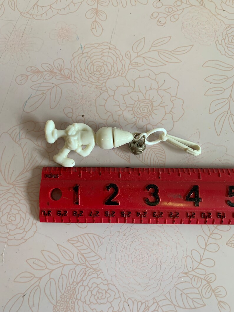 May include: White plastic keychain featuring a cartoon character with a large head and a small body. The character is holding a small object in its hand. The keychain has a metal ring and a plastic clip.