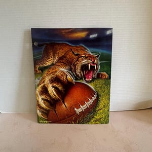 May include: A rectangular three-ring binder cover featuring a graphic illustration of a snarling bobcat with its claws extended over a brown leather football. The scene is set on a green grass field under stadium lights at night.