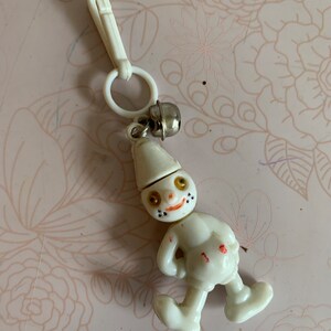 May include: White plastic keychain with a small bell and a cartoon figure of a person with a hat and a smiling face. The figure has black eyes, a red nose, and black dots for a mouth.