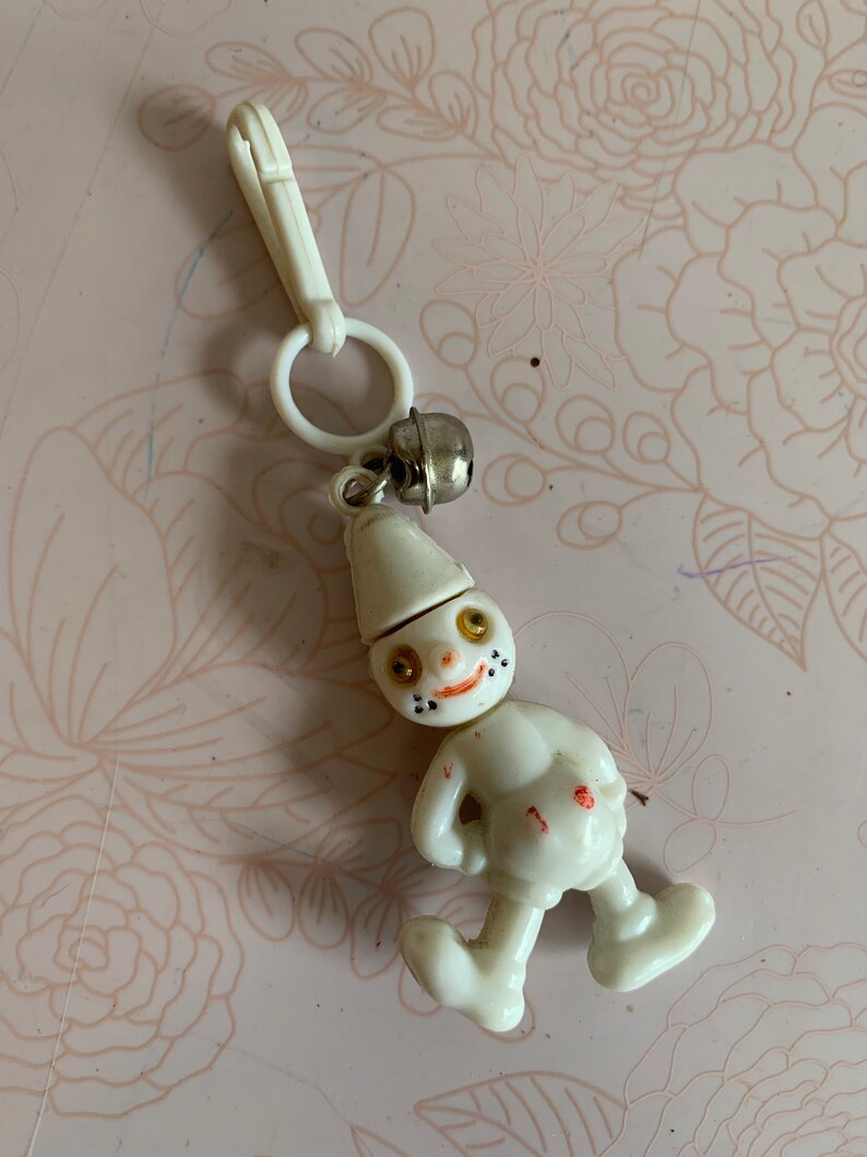 May include: White plastic keychain with a small bell and a cartoon figure of a person with a hat and a smiling face. The figure has black eyes, a red nose, and black dots for a mouth.