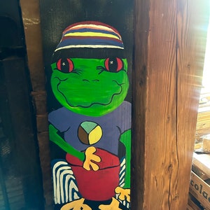 May include: A hand-painted wooden sign featuring a cartoon frog wearing a striped hat, a purple shirt with a peace sign, and red pants. The frog is sitting on a striped stool.