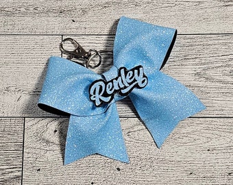Puede incluir: Un lazo de brillantina azul claro con una etiqueta de nombre negra que dice "Renley". El lazo está unido a un llavero plateado.