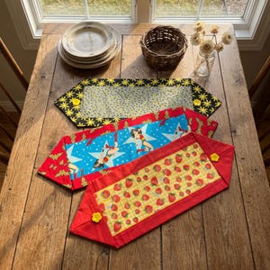 May include: Three rectangular fabric table runners with pointed ends. The top runner is black with yellow star patterns. The middle runner features a blue background with Wonder Woman graphics. The bottom runner has a yellow strawberry print.