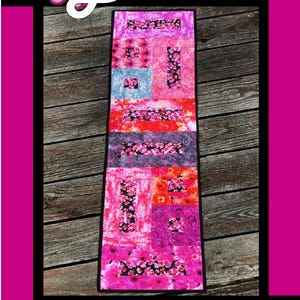 May include: A rectangular table runner featuring a patchwork design of floral and abstract patterns in shades of pink, red, purple, and black. The text Bloom is at the top, and Tricia Lynn Maloney Orphan Quilt Designs for Villa Rosa Designs is at the bottom.