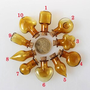 Puede incluir: Diez pequeñas botellas de vidrio ámbar con tapones de corcho. Las botellas son de varias formas, incluyendo redonda, corazón y lágrima. Las botellas están dispuestas en círculo sobre un fondo blanco.