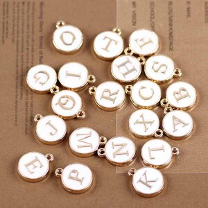 May include: A set of 17 gold-toned metal charms with white enamel letters. Each charm is a circle with a letter printed in gold on the front.
