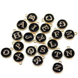 May include: A set of 26 gold-colored alphabet letter charms with black enamel. Each charm features a single letter of the alphabet in a white font.