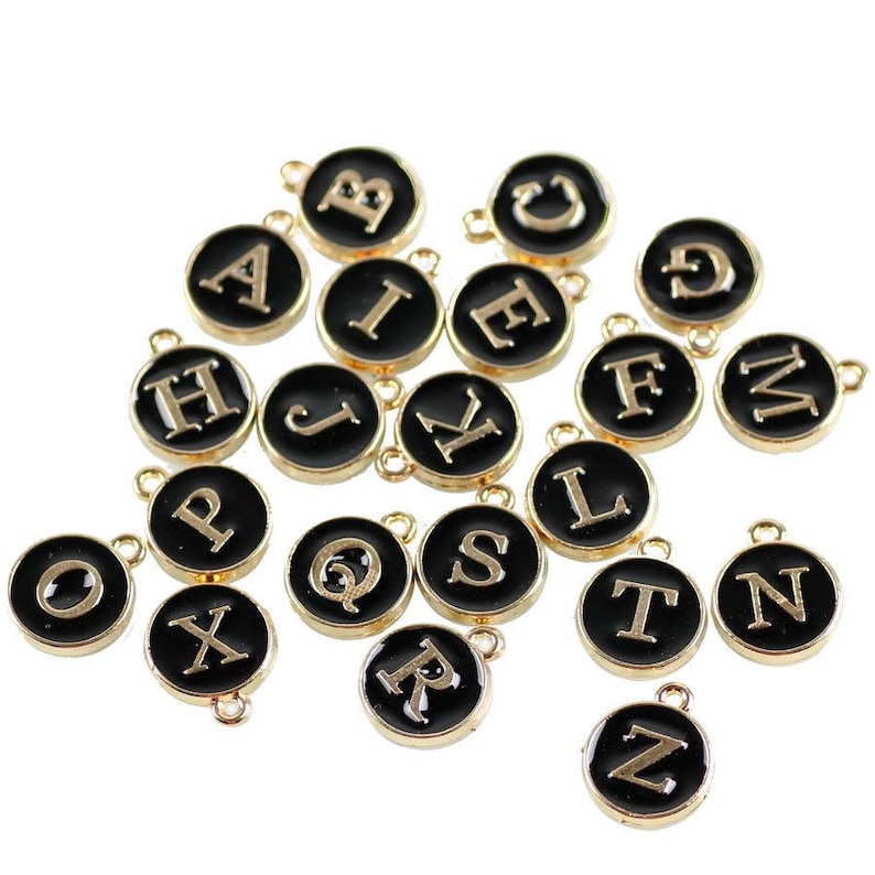 May include: A set of 26 gold-colored alphabet letter charms with black enamel. Each charm features a single letter of the alphabet in a white font.