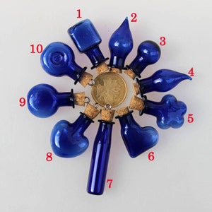 Può includere: Dieci piccole bottiglie di vetro blu con tappi di sughero. Le bottiglie sono di varie forme, tra cui rotonda, a forma di cuore e a forma di lacrima. Le bottiglie sono disposte in cerchio su una superficie bianca.