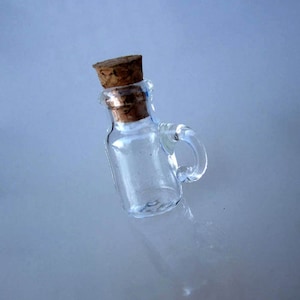 Op de afbeelding: Een klein, helder glazen flesje met een kurkstop en een handvat. De fles is leeg en heeft een ronde bodem.