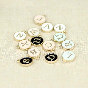 May include: Assortment of gold-tone metal charms with different letters of the alphabet on them. The charms are painted in various colors, including black, white, and pastel.
