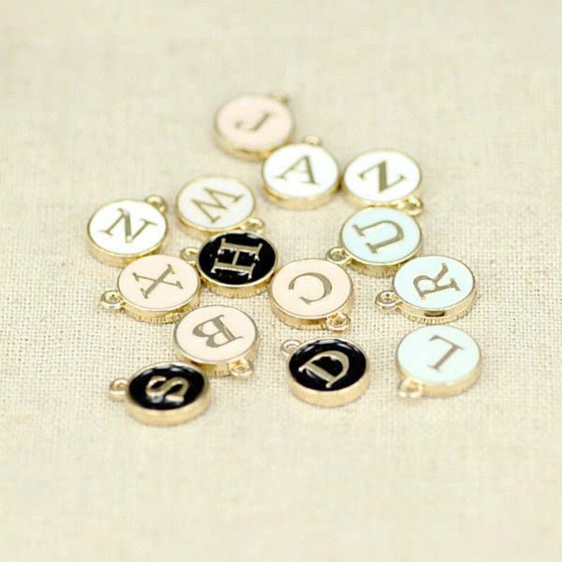 May include: Assortment of gold-tone metal charms with different letters of the alphabet on them. The charms are painted in various colors, including black, white, and pastel.