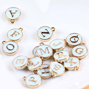 May include: A collection of 15 gold-toned metal charms with white enamel and gold lettering. Each charm features a different letter of the alphabet. The charms are perfect for adding a personalized touch to jewelry, crafts, and other projects.