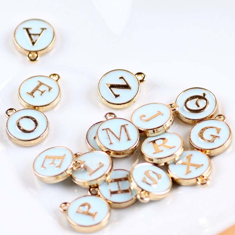 May include: A collection of 15 gold-toned metal charms with white enamel and gold lettering. Each charm features a different letter of the alphabet. The charms are perfect for adding a personalized touch to jewelry, crafts, and other projects.