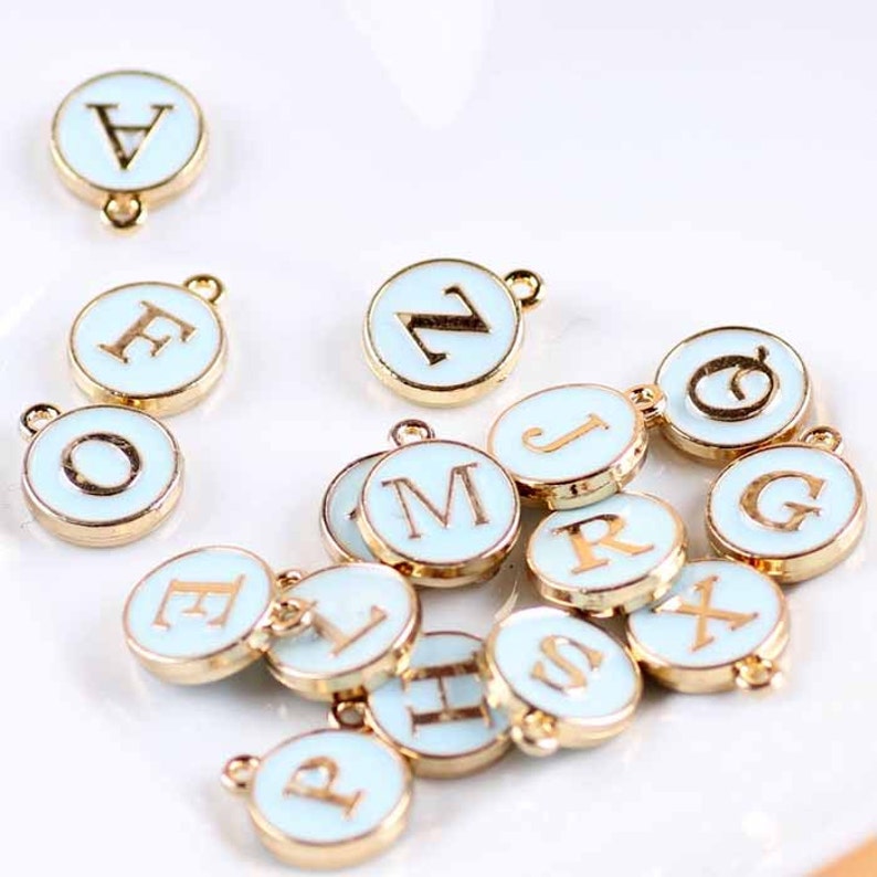 May include: A collection of 15 gold-toned metal charms with a light blue enamel finish. Each charm features a different letter of the alphabet in a gold-toned font.