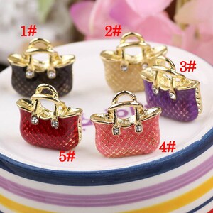 May include: Five gold-tone handbag charms with enamel and rhinestone accents. The charms are in different colors: black, beige, purple, pink, and red.