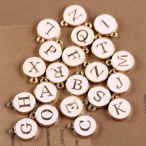 May include: A collection of gold-colored metal letter charms with white enamel. Each charm features a different letter of the alphabet in a gold-colored font.