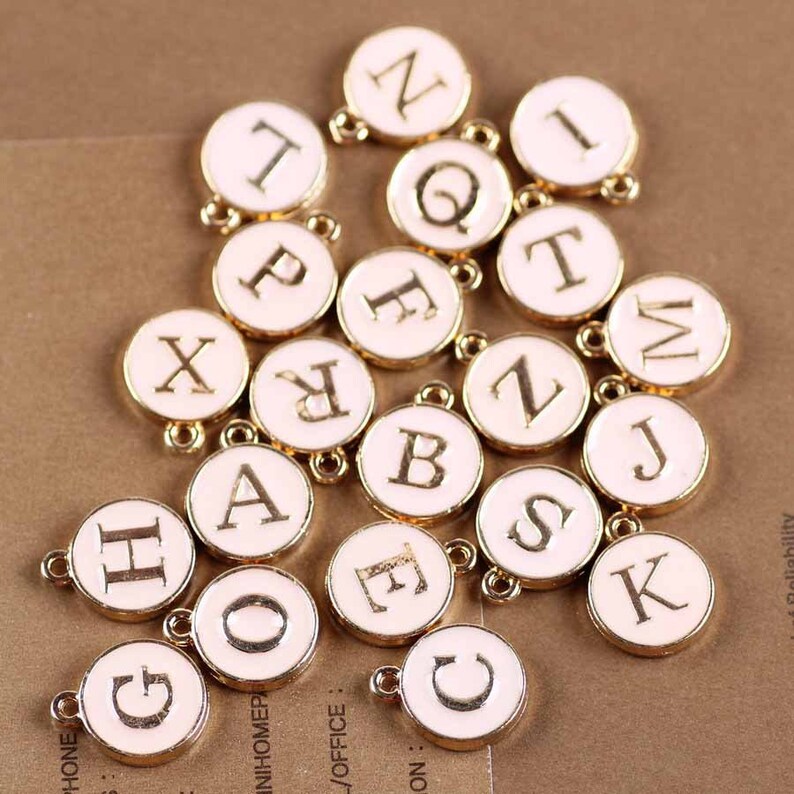 May include: A collection of gold-colored metal letter charms with white enamel. Each charm features a different letter of the alphabet in a gold-colored font.