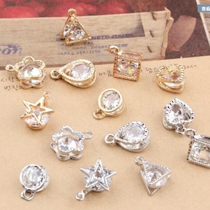 May include: A collection of small, decorative charms in various shapes, including stars, hearts, and teardrops. Each charm features a clear, faceted gemstone set in a gold or silver-toned metal frame. The charms are designed for jewelry making or crafting.
