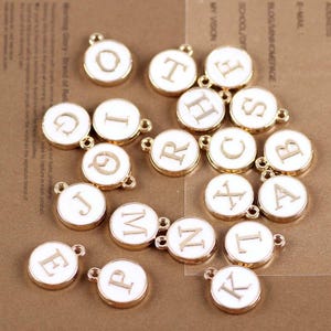 May include: A collection of 18 gold-colored metal charms with white enamel and letters of the alphabet. Each charm has a loop for attaching to a necklace, bracelet, or other jewelry.