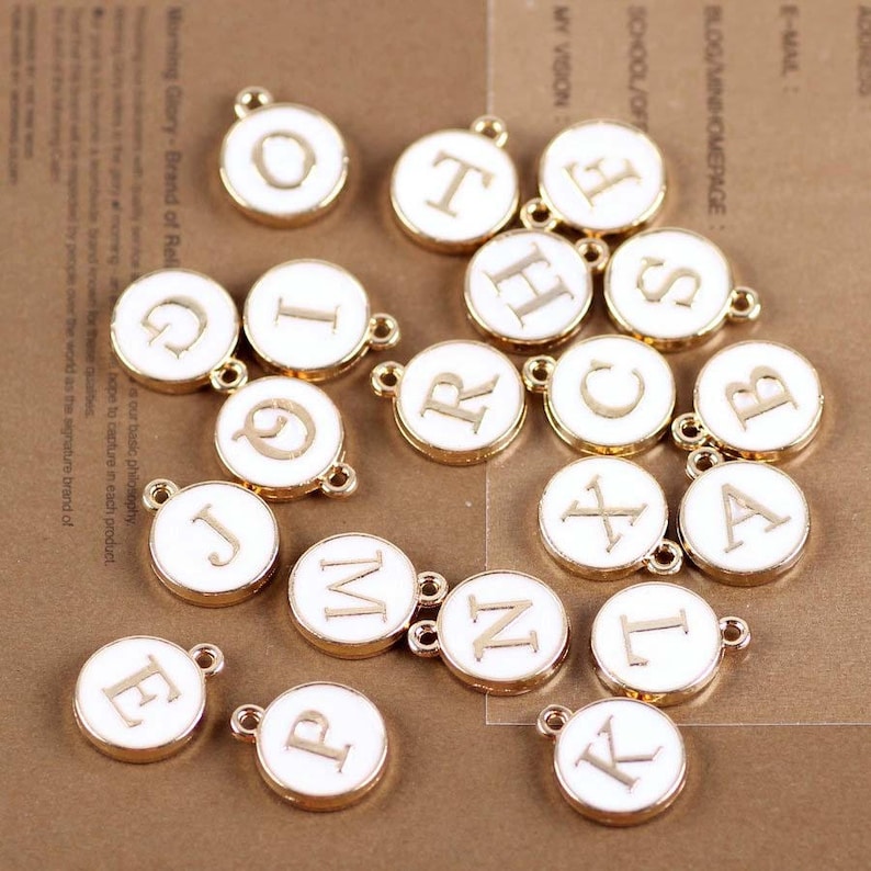 May include: A collection of 18 gold-colored metal charms with white enamel and letters of the alphabet. Each charm has a loop for attaching to a necklace, bracelet, or other jewelry.