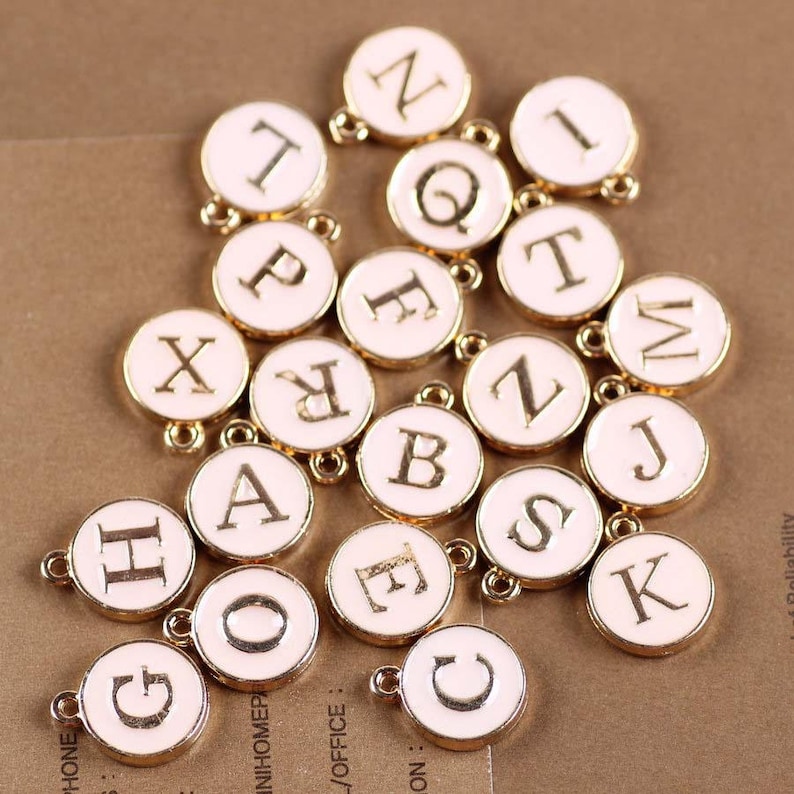 May include: A collection of 20 gold-colored charms with white enamel and letters of the alphabet. The charms are arranged in a random pattern on a brown surface.