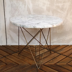 May include: A round side table with a white marble top and a gold-colored metal base. The base has a geometric design with multiple intersecting lines. The table is set on a wooden floor.
