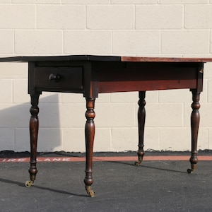 May include: A dark brown antique table with a drawer and turned legs. The table has a rectangular top with a drop-leaf design. The legs are on small caster wheels.