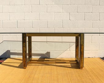 May include: A rectangular coffee table with a clear glass top and a gold-colored metal frame. The table is set against a white brick wall and sits on a light brown surface. The table measures approximately 48 inches long.