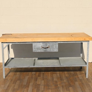 May include: A vintage butcher block table with a light wood top and a metal frame. The table has a drawer with a metal handle and a lower shelf. The table is suitable for a kitchen or workshop.