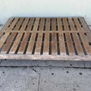 May include: A wooden bed frame with a slatted design. The frame is rectangular with a light brown finish and sits on four short legs. The slats run lengthwise across the frame.
