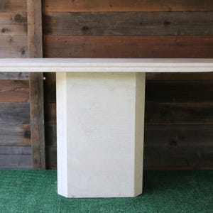 May include: A rectangular, off-white stone table with a geometric pedestal base. The table has a flat top and is set against a wooden plank backdrop and green turf. The table's dimensions are approximately 60 inches long and 20 inches wide.