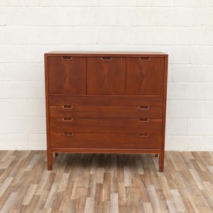 May include: A mid-century modern wooden dresser with a warm brown finish. The dresser has three drawers and four upper cabinet doors. The doors have a diamond-shaped design. The dresser is set on four tapered legs.