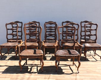 May include: A set of eight brown wooden chairs with woven rush seats. Each chair features a tall, curved ladder-back design with horizontal slats and cabriole legs. The wood has a dark, polished finish with carved floral details on the front.