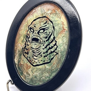 6"x8" black oval plaque with inset resin pour in greens, coppers and ivory with image of The Creature from the Black Lagoon ready for wall hanging.